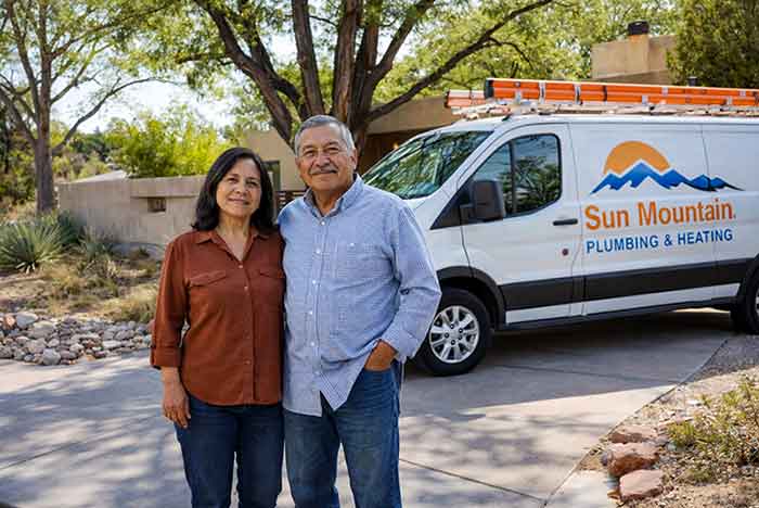 A Hispanic couple in their mid 60s stand in a residential driveway beside a branded Sun Mountain Plumbing & Heating service van, with a Southwestern-style stucco home and mature trees in the background.