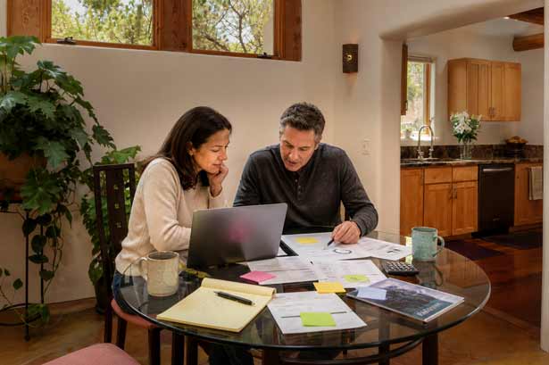 Couple at a kitchen table reviewing documents and notes beside an open laptop
